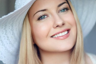 Yaz cilt bakımı ve beyaz dişler, kumsal şapkalı sarışın kadın yumuşak bir şekilde gülümsüyor, açık tenli ve açık renk makyajlı.