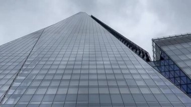 Londra 'daki Çömlek' in dramatik yükseliş manzarası, cam cephesi bulutlu gökyüzüne doğru inceliyor, modern İngiliz mimarisinin yüksekliğini ve zarafetini yakalıyor, yavaş çekim.