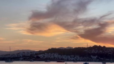 Altın akşam ışığı, Türkiye 'nin Bodrum kentindeki liman ve yamaç kenarındaki evlerin üzerine hafifçe düşüyor. Parlayan bulutların altında demirlemiş tekneler, ağır çekim.