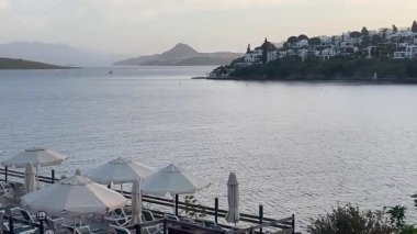Türkiye 'nin Bodrum kenti yakınlarındaki durgun bir koya bakan beyaz şemsiyeler, tepedeki villalar ve akşam gökyüzünün altında yavaşça dinlenen uzak tepeler, yavaş çekim