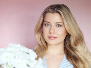 Pastel pembe arkaplanlı yumuşak çiçek cazibesi, soluk mavi elbiseli kadın sakin kadın portresi içinde beyaz kasımpatılar tutuyor.