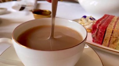 Sütlü taze çay, ince porselenler içinde nazikçe kıvrılmış, zarif sandviçler ve yumuşak çiçekler eşliğinde, sakin bir İngiliz öğleden sonra çayı anında, yavaş çekim.