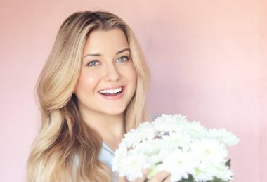 Beyaz kasımpatı tutan, gülümseyen kadın, pastel pembe bir duvara karşı poz veriyor, ışık saçan sıcak yürekli bir İngiliz sıcaklığı ve genç İngiliz cazibesi