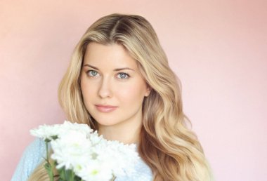 Pastel pembe arkaplanlı yumuşak çiçek cazibesi, soluk mavi elbiseli kadın sakin kadın portresi içinde beyaz kasımpatılar tutuyor.