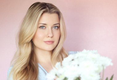 Pastel pembe arkaplanlı yumuşak çiçek cazibesi, soluk mavi elbiseli kadın sakin kadın portresi içinde beyaz kasımpatılar tutuyor.