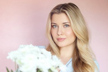 Pastel pembe arkaplanlı yumuşak çiçek cazibesi, soluk mavi elbiseli kadın sakin kadın portresi içinde beyaz kasımpatılar tutuyor.