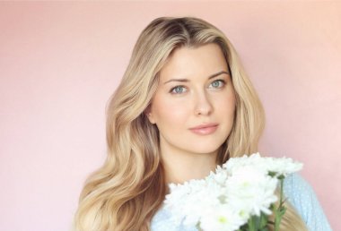 Pastel pembe arkaplanlı yumuşak çiçek cazibesi, soluk mavi elbiseli kadın sakin kadın portresi içinde beyaz kasımpatılar tutuyor.