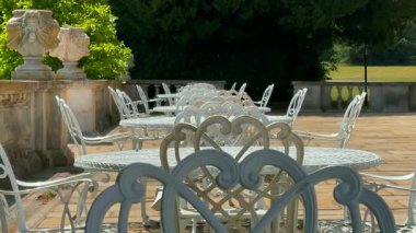 Süslü demir sandalyeler ve masalar bir İngiliz kır köşkünde güneşli teras boyunca dizilmiş, çaydan önce sessizlik, yavaş çekim