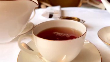 Sütlü taze çay, ince porselenler içinde nazikçe kıvrılmış, zarif sandviçler ve yumuşak çiçekler eşliğinde, sakin bir İngiliz öğleden sonra çayı anında, yavaş çekim.