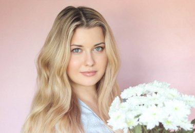 Pastel pembe arkaplanlı yumuşak çiçek cazibesi, soluk mavi elbiseli kadın sakin kadın portresi içinde beyaz kasımpatılar tutuyor.