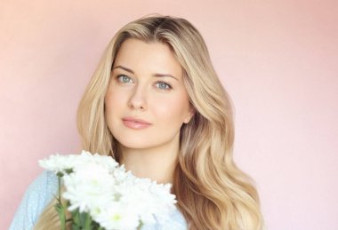 Pastel pembe arkaplanlı yumuşak çiçek cazibesi, soluk mavi elbiseli kadın sakin kadın portresi içinde beyaz kasımpatılar tutuyor.