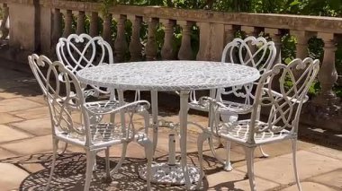 Süslü beyaz işlemeli demir sandalyeler bir bahçe masasının etrafında, taş korkulukların yanında, yumuşak güneş ışığı ile benekli, yavaş çekim