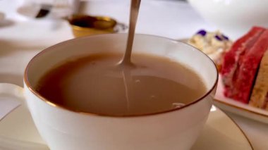 Sütlü taze çay, ince porselenler içinde nazikçe kıvrılmış, zarif sandviçler ve yumuşak çiçekler eşliğinde, sakin bir İngiliz öğleden sonra çayı anında, yavaş çekim.