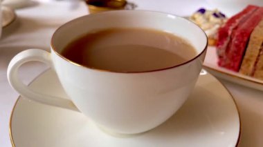 Sütlü taze çay, ince porselenler içinde nazikçe kıvrılmış, zarif sandviçler ve yumuşak çiçekler eşliğinde, sakin bir İngiliz öğleden sonra çayı anında, yavaş çekim.