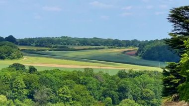Yumuşak mavi gökyüzünün altındaki katmanlı İngiliz kırsal tepelerinin ve ağaçların geniş bir görüntüsü, yaz ışığı altında, yavaş çekimde.