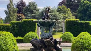 Süslü İngiliz çeşmesi kırpılmış çalı ve yontulmuş çitlerle çerçevelenmiş, ötesinde resmi bahçe ağaçları, zarif kırsal malikane düzeni, yavaş çekim