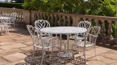 Süslü beyaz işlemeli demir sandalyeler bir bahçe masasının etrafında, taş korkulukların yanında, yumuşak güneş ışığı ile benekli, yavaş çekim