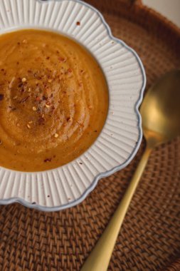 Creamy bowl of pumpkin or butternut squash soup topped with cracked pepper and chilli flakes, placed beside golden spoon on woven mat, concept of homemade seasonal food in the English kitchen