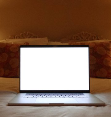 Laptop with a blank white screen glowing in a dim bedroom. Technology, work from home, and leisure concepts. Mockup copyspace