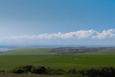 Seven Sisters cliffs appearing in the distance beyond green fields and a distant cloudy sky. East Sussex countryside, England