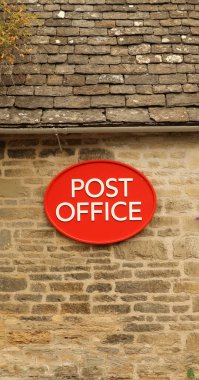 Post office red oval sign with white text fixed on an old rustic stone wall, showing traditional building architecture and mail service concept. Cotswolds, England