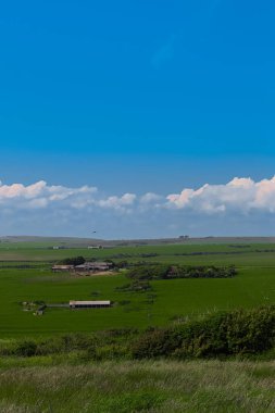 Parlak mavi gökyüzü altında tarım yapıları ile kırsal alanı kaplayan geniş yeşil alanlar. Doğu Sussex kırsal bölgesi, İngiltere