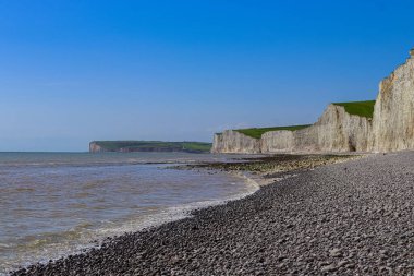 Seven Sisters white chalk cliffs standing over a pebble beach and the vibrant blue English Channel, showcasing the stunning coastal landscape. East Sussex, England