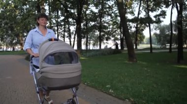 Bebek arabasında bir bebekle anne parkta yol boyunca yavaşça yürür. 4K Ağır çekim
