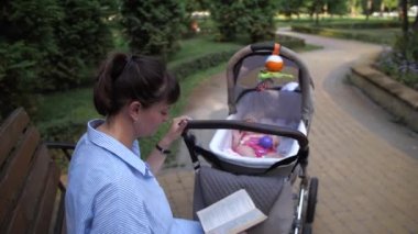 Genç bir anne tek eliyle bebeği bebek arabasında sallıyor. Kız parkta bir bankta oturmuş kitap okuyor. 4K Ağır çekim