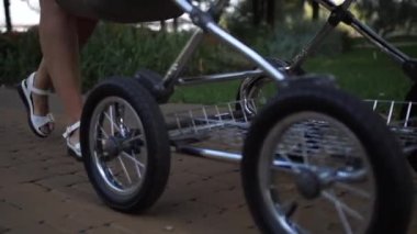 Yakın plan. Bebek arabasından tekerlekler geliyor, onu kadın bacakları takip ediyor. Annem bebekle yürüyor..