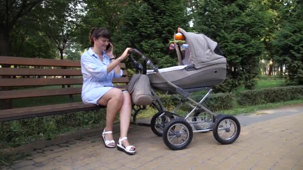 Egy Fiatal Anya Padon Rázza Babakocsit Miközben Telefonál Kamera Mozgásban — Stock videók