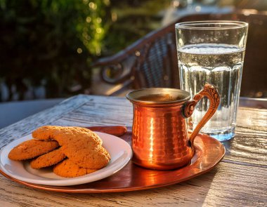 Enjoy a delightful moment with traditional Greek coffee served in a copper pot, accompanied by sweet cookies and refreshing cold water on a charming outdoor table. clipart