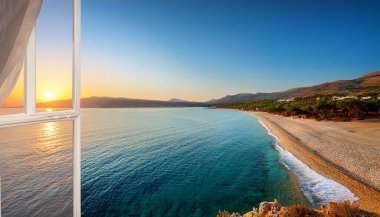 Bright colors flood the sky as the sun sets over a picturesque Greek beach. Waves gently lap at the sandy shore as the tranquil waters reflect the evening light. Lush green hills frame the landscape, inviting viewers to relax and enjoy the natural be clipart