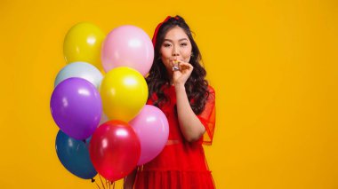 asian woman in red dress holding colorful balloons and blowing party horn isolated on yellow  clipart