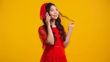 happy asian woman in red dress talking on smartphone while twirling hair isolated on yellow  clipart
