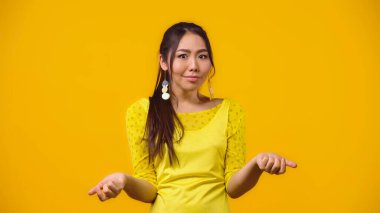 confused asian woman showing shrug gesture isolated on yellow clipart