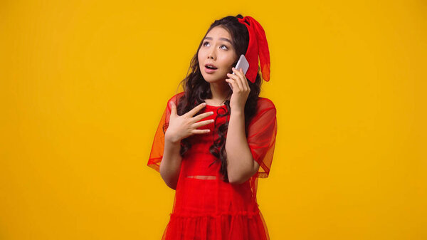 emotional asian woman in red dress talking on smartphone isolated on yellow 
