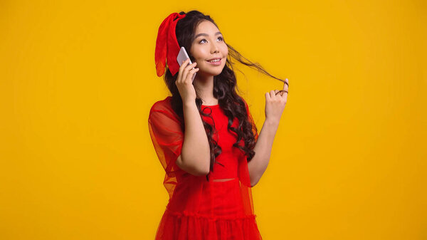 happy asian woman in red dress talking on smartphone while twirling hair isolated on yellow 