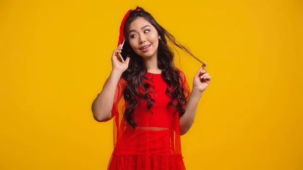 stock image cheerful asian woman in red dress talking on smartphone while twirling hair isolated on yellow 