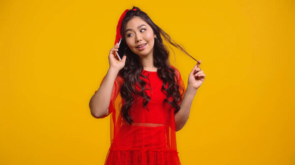 cheerful asian woman in red dress talking on smartphone while twirling hair isolated on yellow 