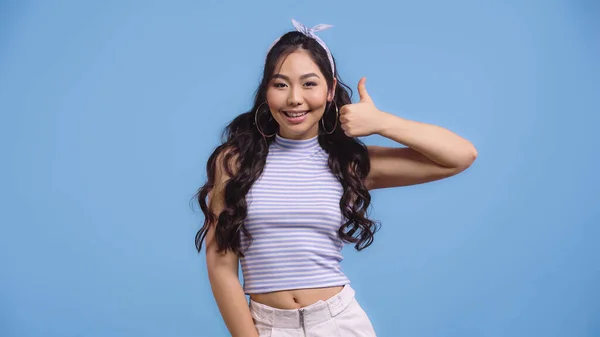 happy asian woman in hoop earrings showing thumb up isolated on blue