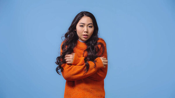 brunette and young asian woman in sweater feeling cold isolated on blue