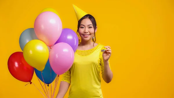 幸せアジアの女性でお祝いのキャップ保持カラフルな風船とパーティーホーン孤立で黄色 ストック画像