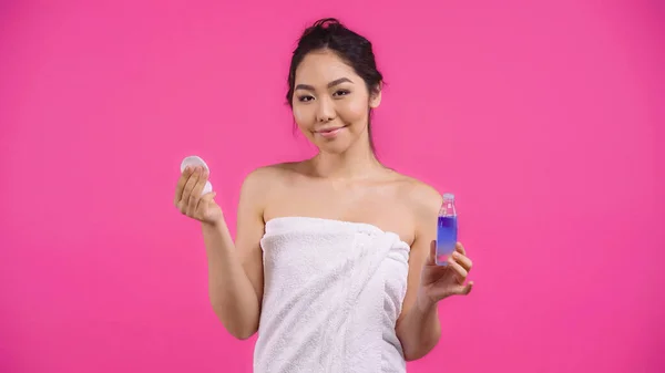 Asian young woman in towel holding cotton pad and micellar water isolated on pink — Stock Photo