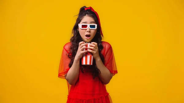 Conmocionado mujer asiática en 3d gafas sosteniendo palomitas de maíz y viendo aterrador película aislado en amarillo - foto de stock