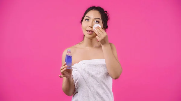 Asiatico giovani donna in asciugamano tenendo cotone pad con micellare acqua mentre pulizia faccia isolato su rosa — Foto stock