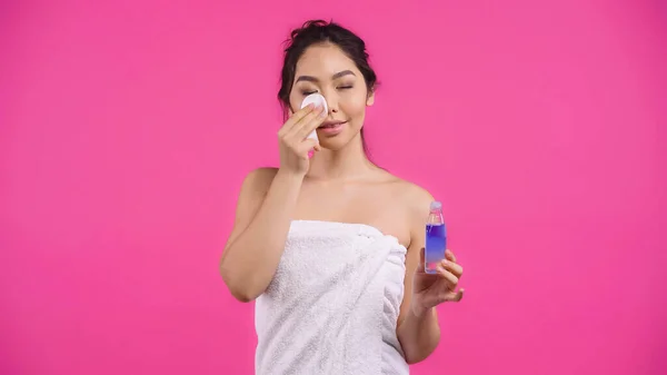 Young asian woman in towel holding cotton pad with micellar water while cleansing face isolated on pink — Stock Photo