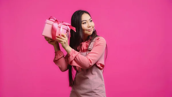 Glücklich junge asiatische Frau schüttelt eingewickelt Geschenk isoliert auf rosa — Stockfoto