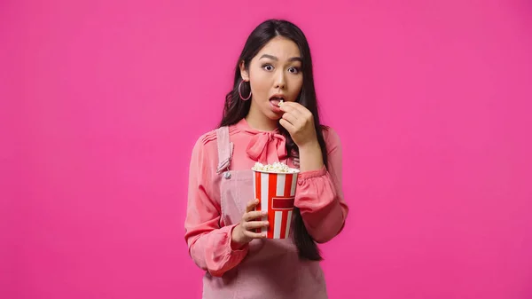 Young asian woman with opened mouth eating salty popcorn bucket isolated on pink — Stock Photo