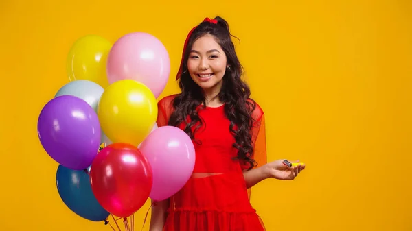 Alegre asiático mujer en rojo vestido celebración colorido globos y partido cuerno aislado en amarillo - foto de stock
